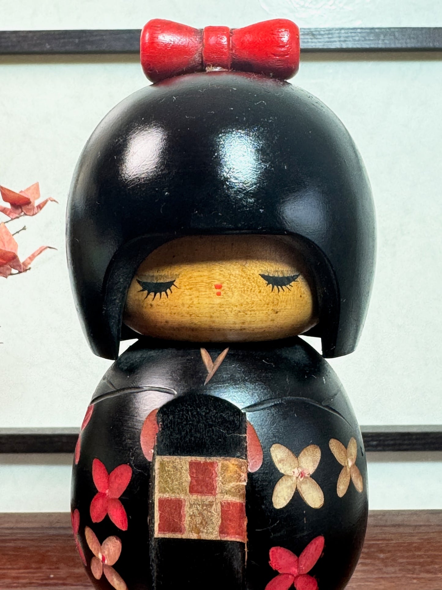 kokeshi de Miyashita Hajime, noire avec fleurs cerisier blanches et rouge, noeud rouge sur la tête, gros plan sur son visage en bois clair, souriant