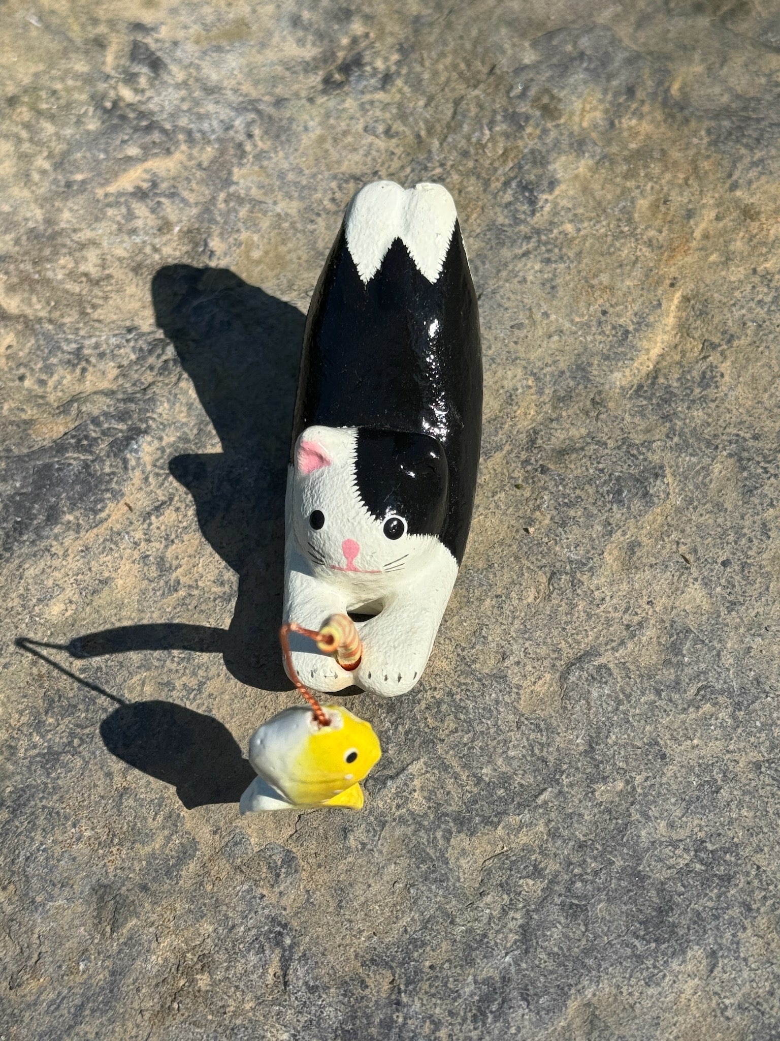 figurine japonaise, chat noir et blanc, allongé sur le ventre les pattes arriere en l'air, tenant une canne à pêche sur laquelle est suspendu un poisson, en papier maché, vu de dessus