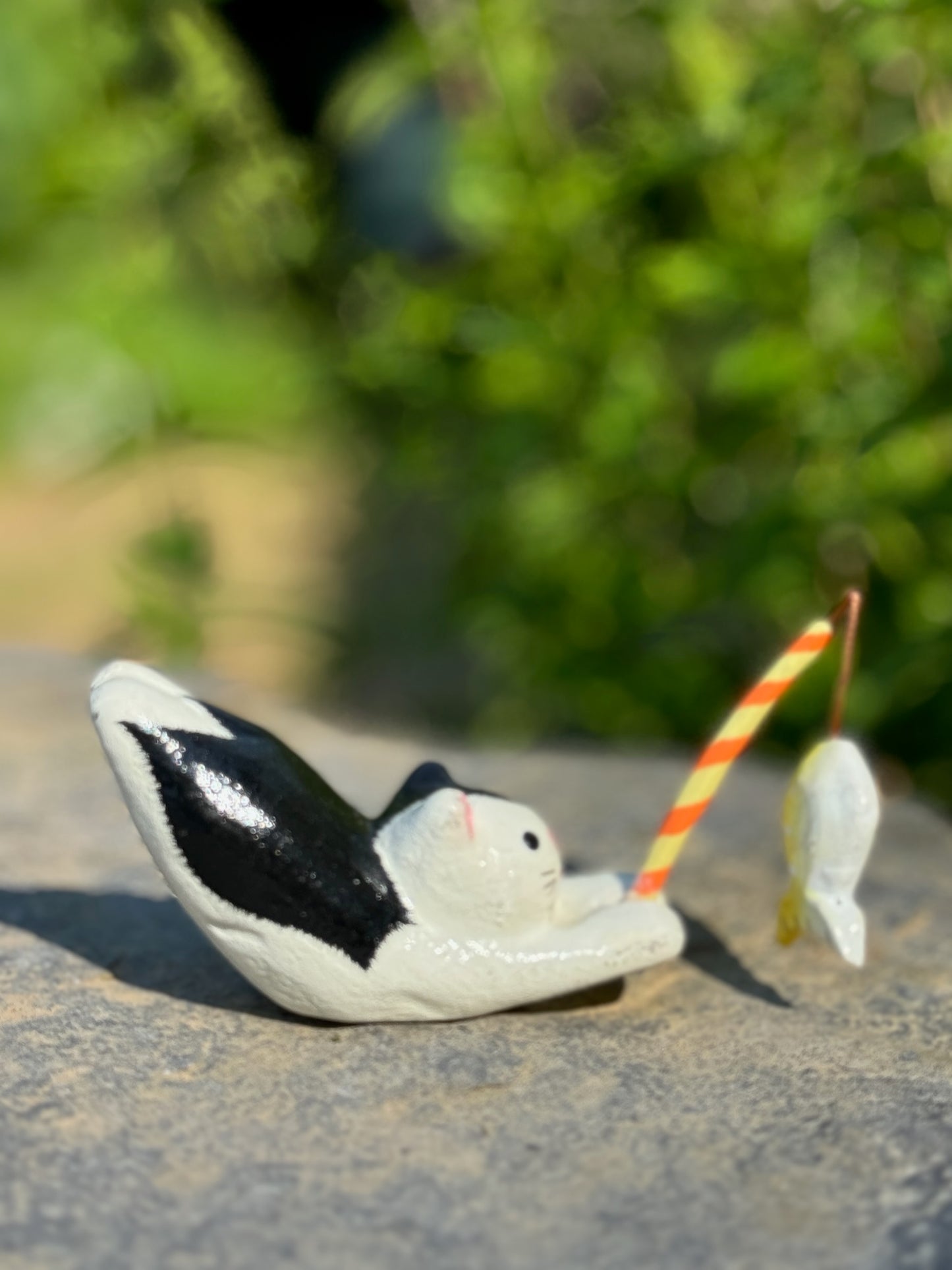 figurine japonaise, chat noir et blanc, allongé sur le ventre les pattes arriere en l'air, tenant une canne à pêche sur laquelle est suspendu un poisson, en papier maché