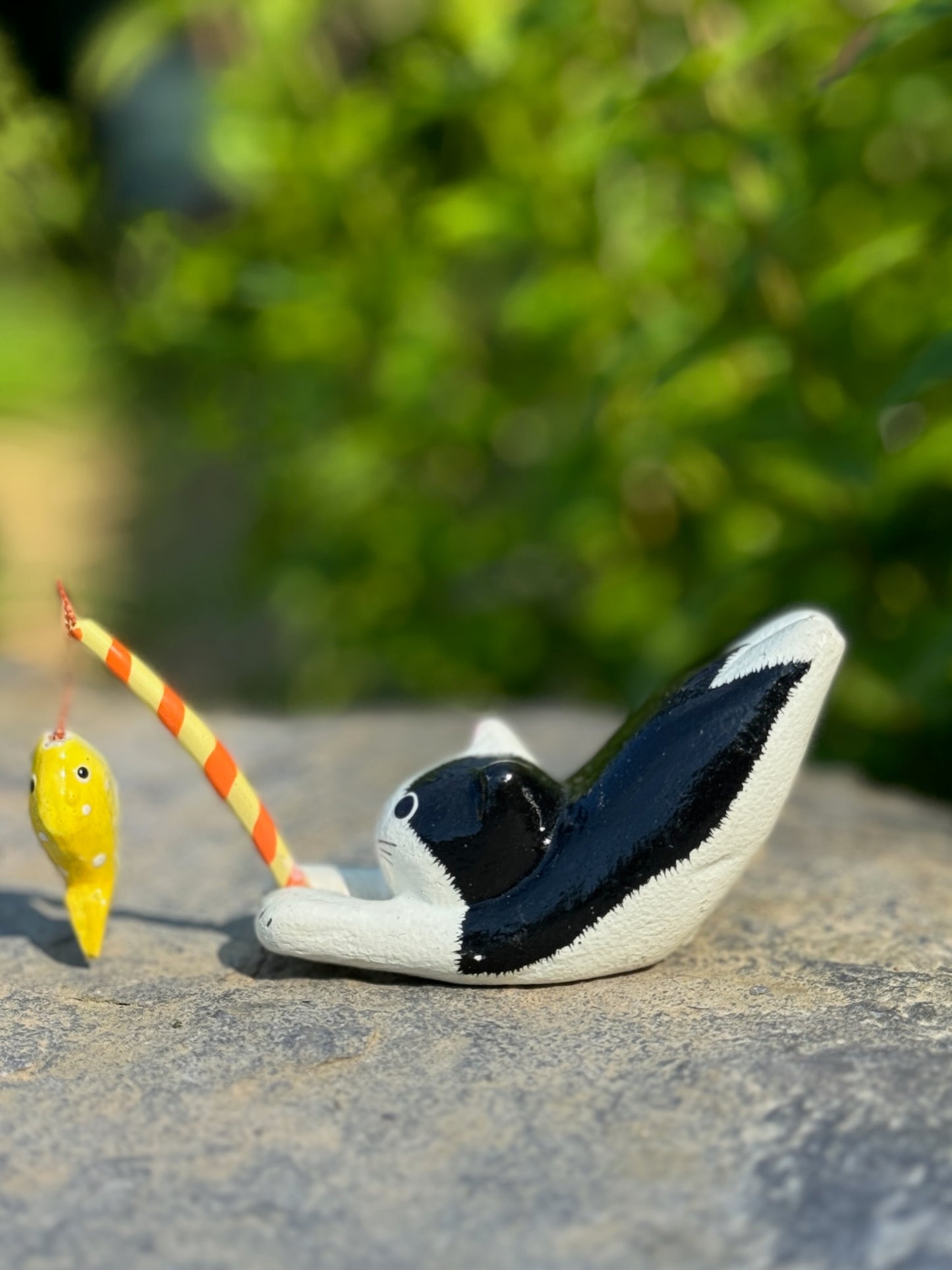 figurine japonaise, chat noir et blanc, allongé sur le ventre les pattes arriere en l'air, tenant une canne à pêche sur laquelle est suspendu un poisson, en papier maché, profil gauche