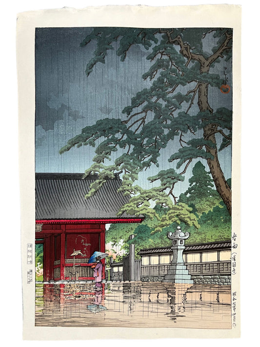 estampe japonaisee de Kawase Hasui, pluie sur le temple rouge de Gokokkuji, personne sous parapluie bleu, lanterne en pierre, pin