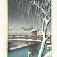 estampe japonaise de Kawase Hasui, deux femmes avec parapluies traverse un pont japonais sous une tempête de neige, le soir, en premier plan un arbre enneigé, au loin un village élairé