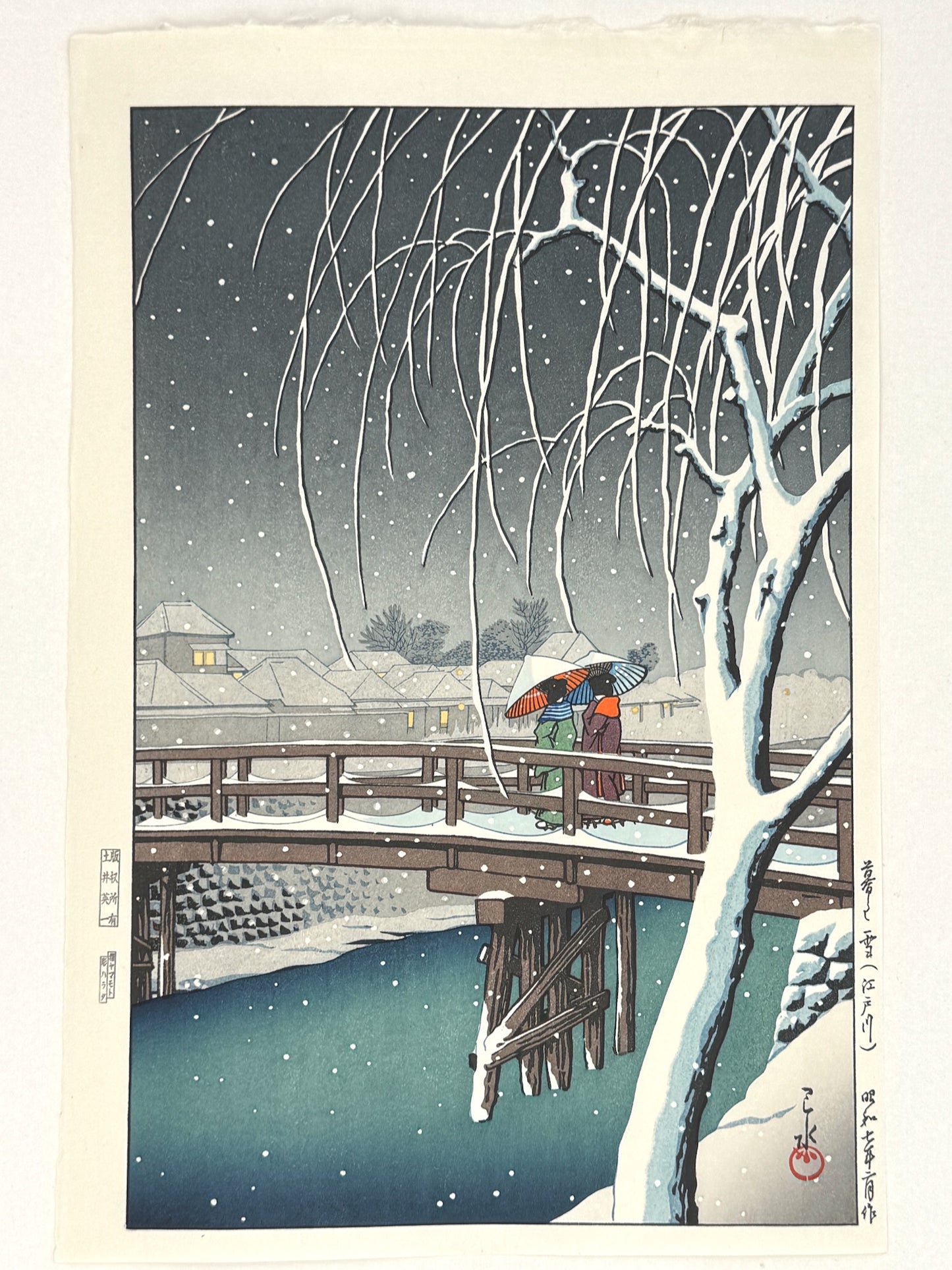 estampe japonaise de Kawase Hasui, deux femmes avec parapluies traverse un pont japonais sous une tempête de neige, le soir, en premier plan un arbre enneigé, au loin un village élairé