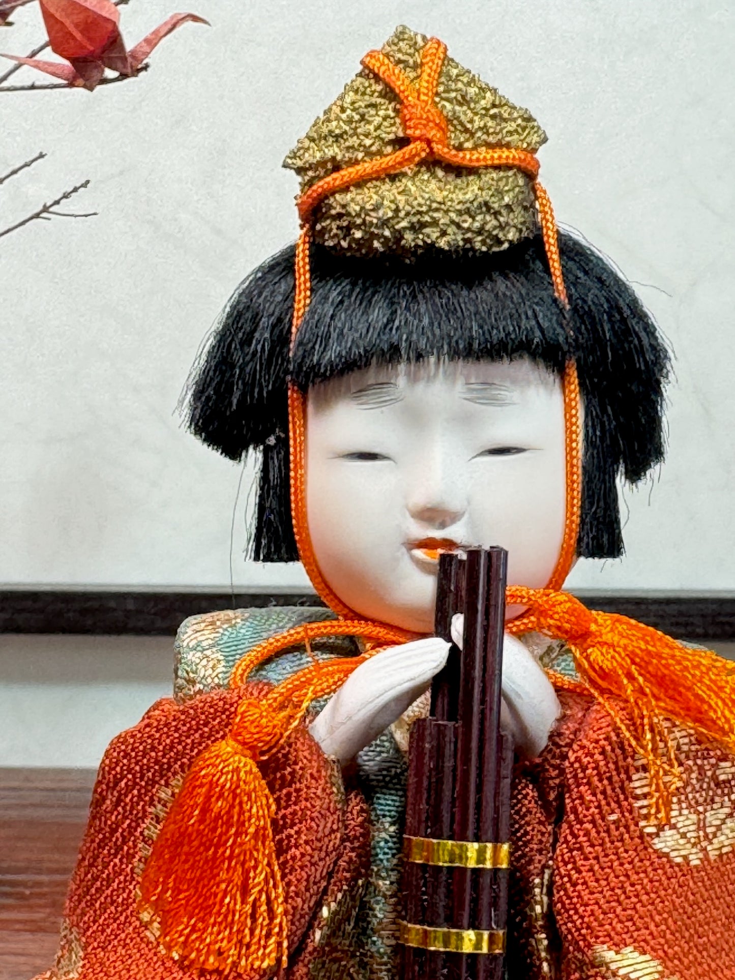 poupée japonaise kimekomi gosho avec un petit chapeau et jouant du sho, orgue à bouhe. Vêtu d'un kimono orande à motif doré, gros plan sur le visage blanc et la flûte