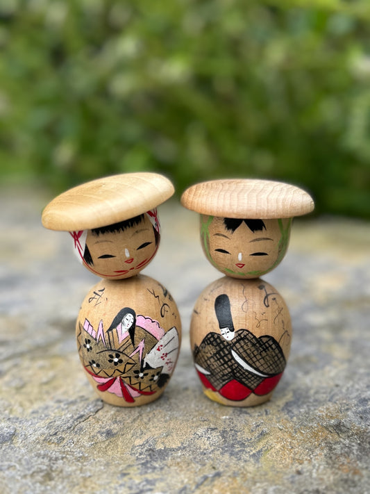 Couple de poupées kokeshi en bois représentant un empereur et une impératrice avec des chapeaux ronds, décorées de motifs traditionnels peints à la main.