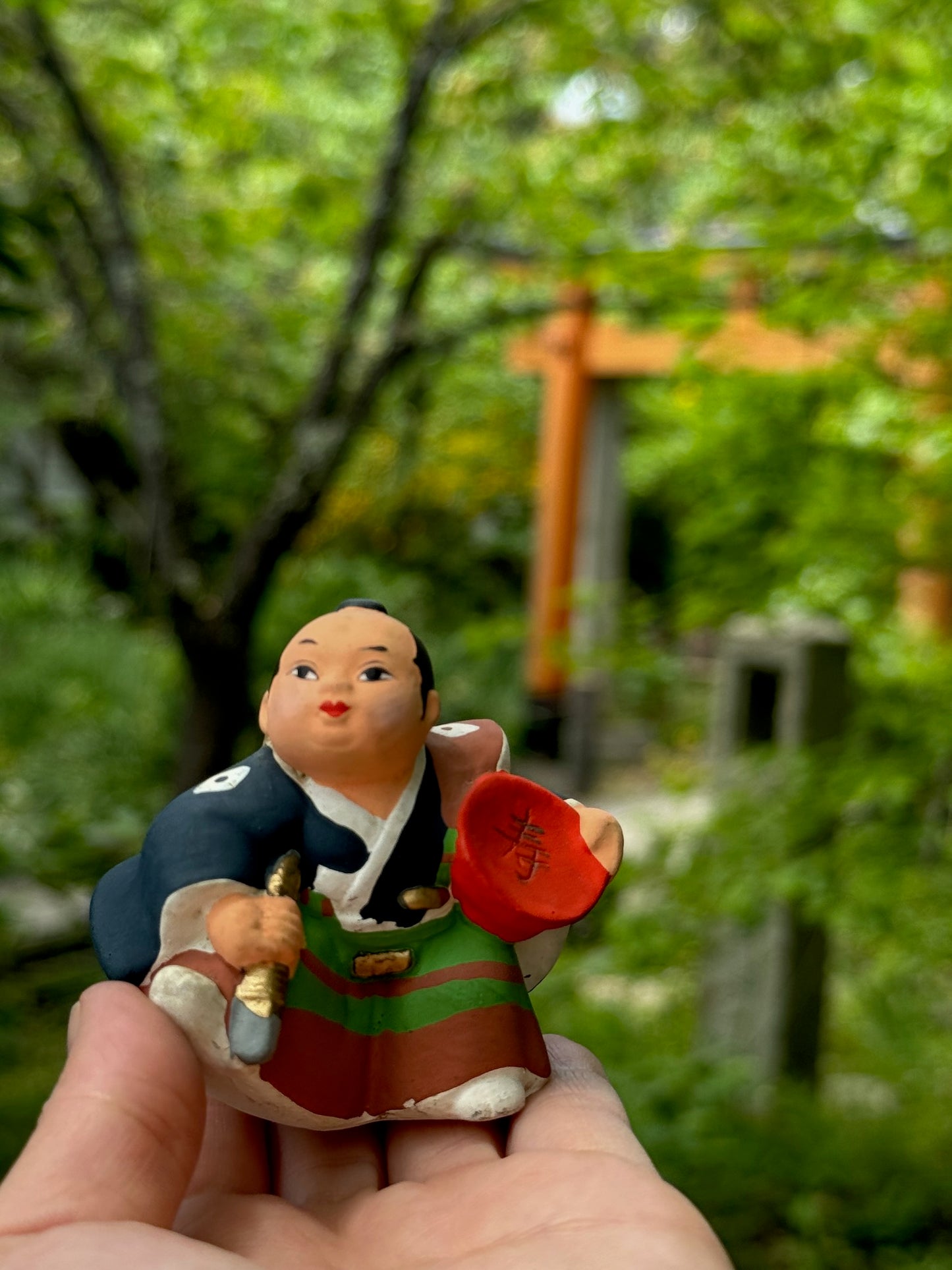 Poupée Samourai en porcelaine biscuit d'Hakata, coupe de sake rouge sous le bras et dague, kimono bleu et hakama vert et marron.